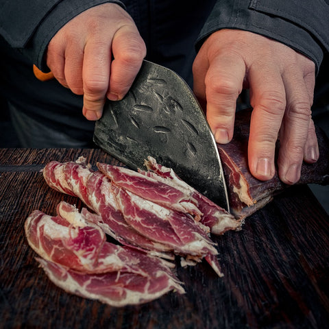Handgeschmiedetes Chef-Messer aus Carbonstahl schneidet Fleisch auf Holzbrett, ideal für Küche und Outdoor-Einsatz.