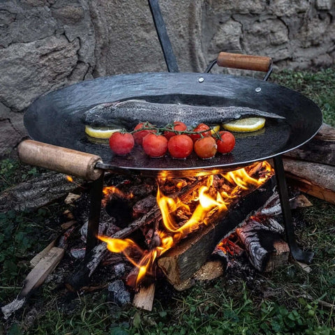 Handgeschmiedete Lagerfeuerpfanne über offenem Feuer mit frischem Fisch und Tomaten, ideal für Outdoor-Kochen.